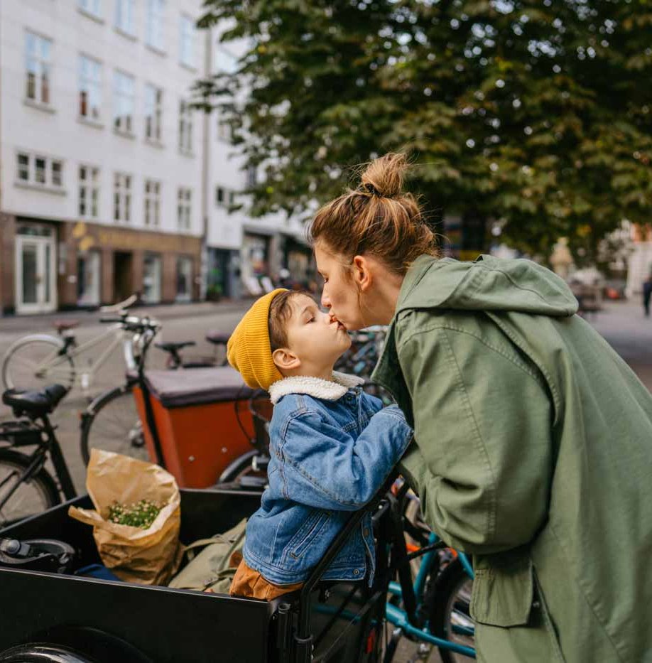 Eine Mutter küsst ihren Sohn der in einem Lastenrad steht.