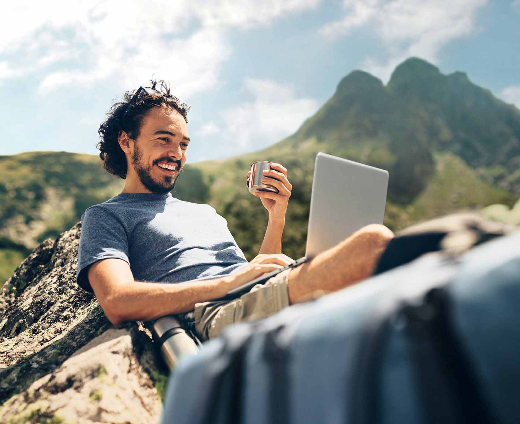 Ein junger Mann sitzt mit seinem Laptop auf einem Berggipfel und prüft mit einem Lächeln seine Geldanlage